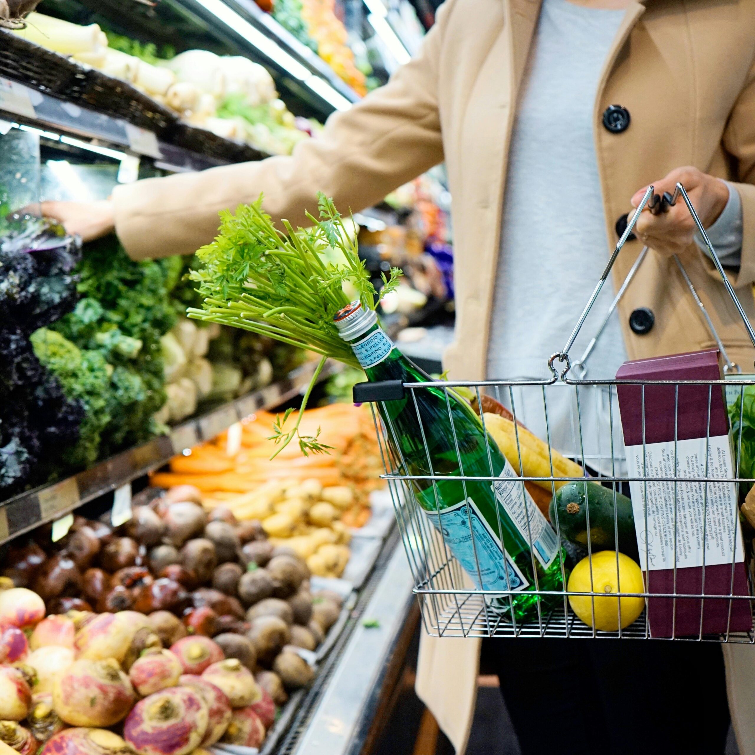 Woman selecting fresh produce in a supermarket with a basket full of groceries.