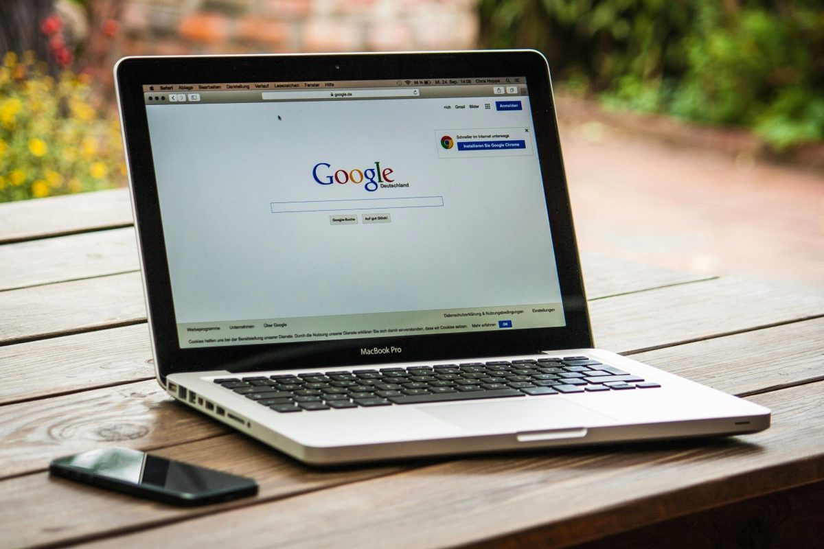 A MacBook Pro displaying Google Search on a wooden table outdoors, next to a smartphone.